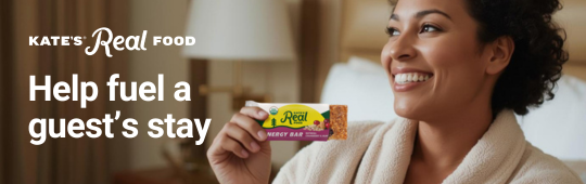 Smiling woman in a bathrobe holding a Kate's Real Food energy bar with text "Help fuel a guest’s stay". Smiling woman in a bathrobe holding a Kate's Real Food energy bar with text "Help fuel a guest’s stay".