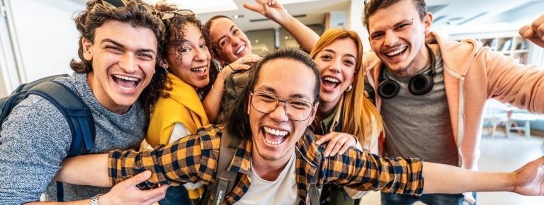 Diverse group of six young adults huddled together indoors, smiling broadly and conveying happiness.